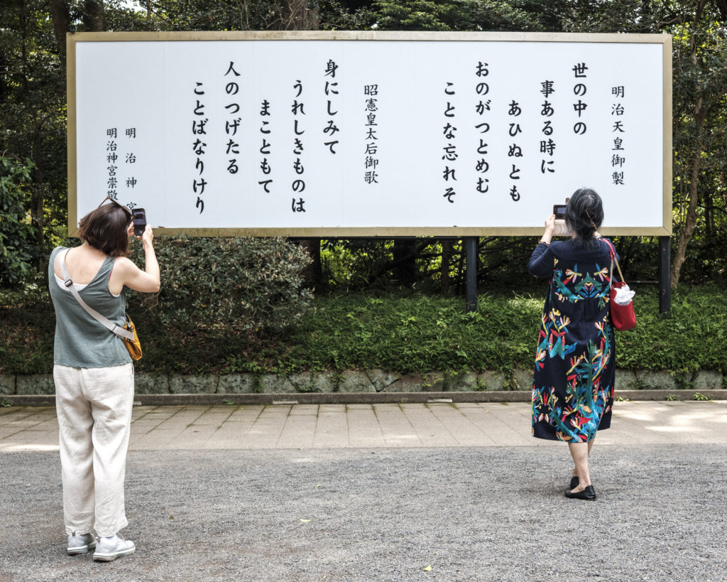 Two women use their smartphones to translate Japanese script written on a wide white sign in the grounds of Meiji Jingu shrine, Tokyo.