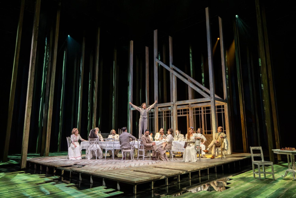 The cast of Summerfolk sitting for an al-fresco dinner outside a deconstructed dacha set that also appears to be a raft. One of the cast, Vlass, is standing on the table shouting with his fists raised.